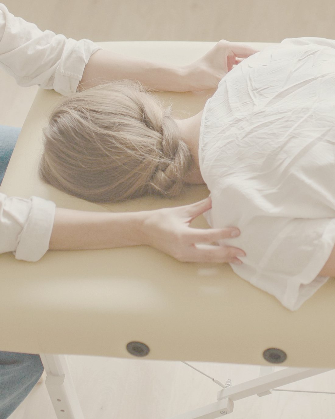 Craniosacrale Behandlung: eine Frau liegt im weißen Hemd auf dem Bauch, die Hände der Therapeutin ruhen behutsam an ihren Schultern